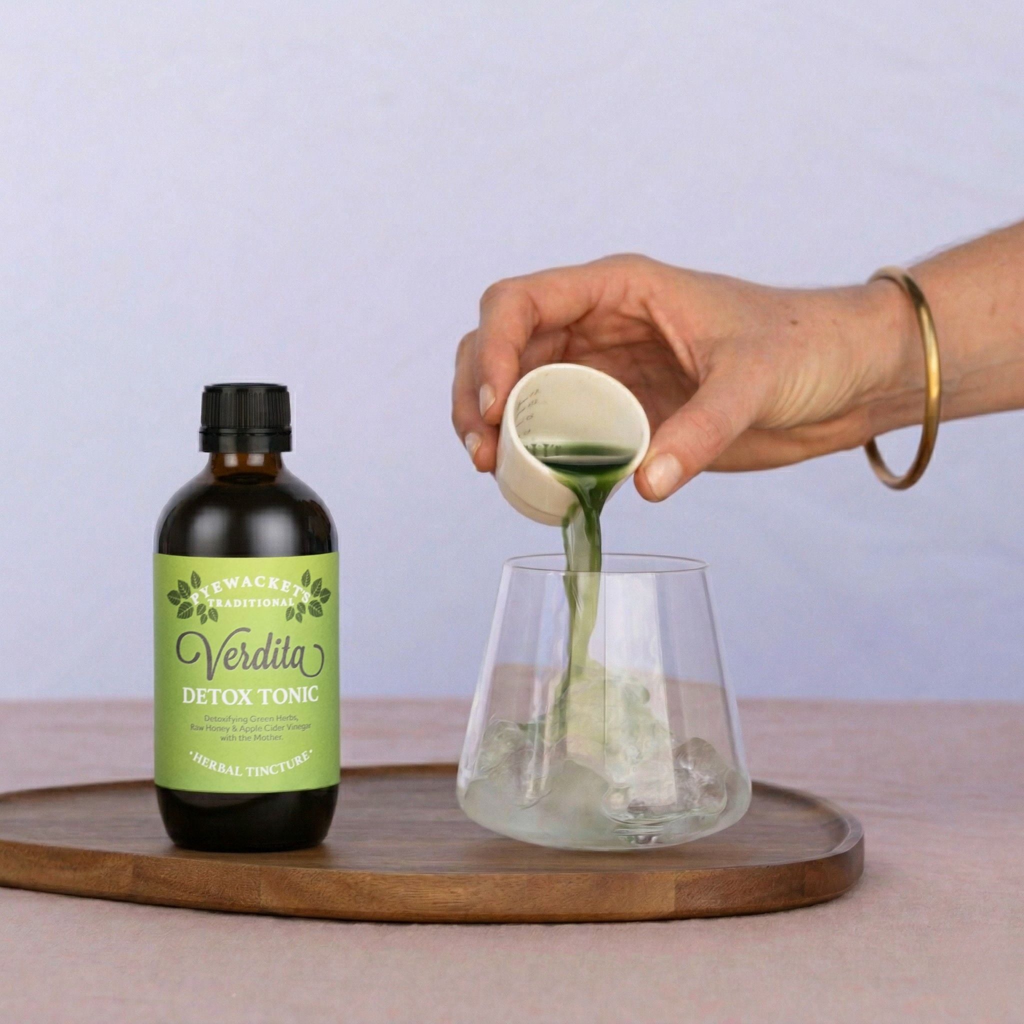 Verdita Detox Tonic being poured from a ceramic measuring cup into a glass on a wooden tray with a pale background.