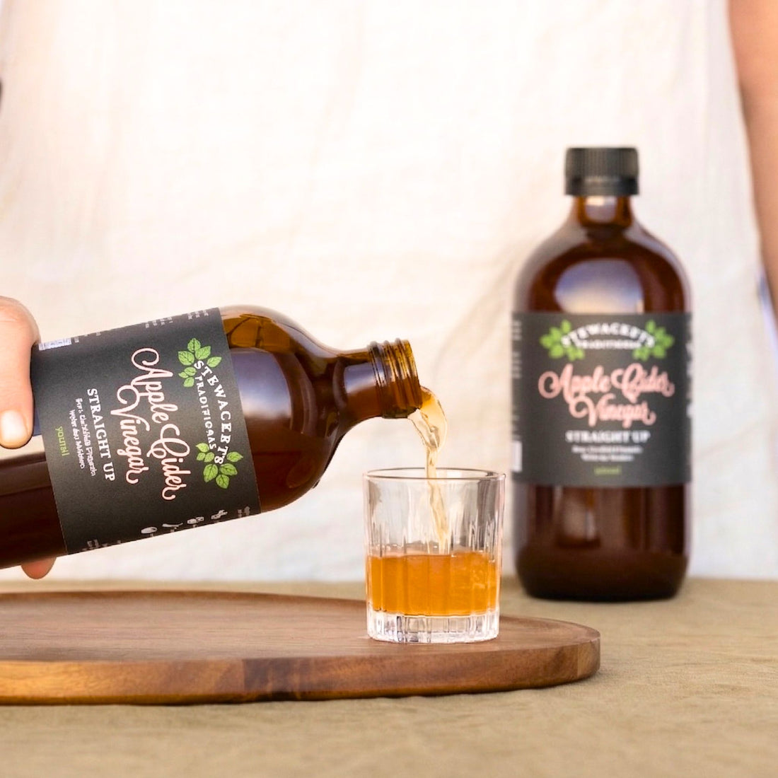 Person pouring apple cider vinegar into a glass with a second bottle in the background.