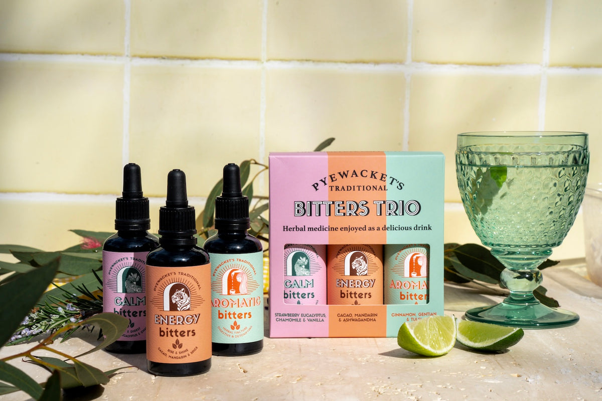 Three dropper bottles of Prewackets Traditional Bitters alongside their colourful gift box, styled with lime slices and a green glass on a tiled surface.